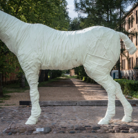 A large statue of a horse wrapped entirely in a white material stands on a gravel walkway, inviting interest from passersby. The background features trees, two historic brick buildings resembling those of Daugavpils Fortress, and a small garden. The pathway extends into the distance under a partly cloudy sky.
