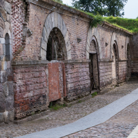 The Daugavpils fortress boasts a historical wall of weathered brick and stone. Arched doorways, partially open, lead into shadowed entries. A cobblestone path, edged by a narrow concrete strip, guides visitors while lush greenery crowns the structure.