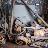 A collection of rusty tools and metal parts is scattered on a floor against a speckled wall in a forgotten corner of the Daugavpils fortress. Items include shovels, pipes, gears, and a radiator. A stone and two small rusted cans are also visible, along with two bricks and a porcelain pitcher.