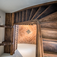 A curved wooden staircase with dark brown steps leads down to a lower floor, reminiscent of the architectural elegance found in Daugavpils Fortress. The ceiling above features an intricate exposed red brick pattern with a central diamond detail. Wall-mounted cylindrical lights cast their glow onto the wooden surfaces.