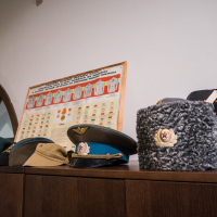 A display of various military hats is arranged on a wooden shelf, reminiscent of those found at Daugavpils Fortress. The collection includes a blue cap with a gold emblem, a beige cap, and a gray fur hat with a badge. Behind them, a framed chart of uniforms is mounted on the wall beneath an exit sign above the wooden archway.
