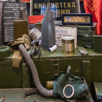 A cluttered display of vintage military items on a green chest evokes memories of Daugavpils' fortress history. Includes a gas mask, hose, tin cans, an old radio, green helmet, and signs with Cyrillic text. A red flag drapes over the side, adding color to the assortment of wartime memorabilia.