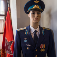 A mannequin dressed as a Soviet military officer stands indoors at the Daugavpils Fortress. The uniform is dark blue with gold accents, adorned with medals and insignia. A red flag with a large star and Cyrillic text is positioned beside it. Sunlight streams through a nearby window, creating a warm ambiance.