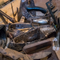 A collection of rusty vintage tools and irons is piled in a wooden crate, nestled within the historic walls of Daugavpils Fortress. The items, with their metal and wooden components, show wear and corrosion, telling tales of age and previous use in this storied setting.
