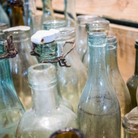 A collection of vintage glass bottles, reminiscent of relics from a forgotten fortress, is displayed on wooden shelves. Some are blue or clear, while others exhibit a cloudy or tinted patina. A few have corks or metal closures, and the surfaces show signs of age, with dust and rustic charm akin to Daugavpils history.