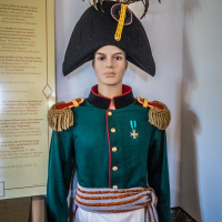 A mannequin dressed in a historical military uniform stands proudly in the Daugavpils fortress exhibition. The outfit includes a dark green jacket with gold epaulettes, a green medal, and a braided belt. A tall black bicorne hat adorned with decorative feathers completes the look. Behind it is a display board with text.