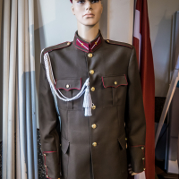 A mannequin dressed in a military uniform with a brown jacket featuring red piping, gold buttons, and an epaulet with a white aiguillette stands proudly in front of the partially visible Latvian flag. Behind it lies the historic Daugavpils Fortress, its presence enhancing this evocative display.
