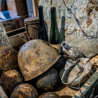 A collection of old, rusted wartime artifacts, including helmets, balls, and a circular metal sign with faded text, hints at the history of Daugavpils Fortress. Two empty green bottles and a damaged metal pitcher add to the military atmosphere in a weathered wooden crate.