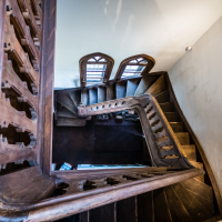 A spiral staircase made of wood is viewed from above, reminiscent of the elegance found in Daugavpils fortress. The stairs curve gracefully, with intricate detailing on the banister. Three decorative windows are visible at the bottom, casting light on the dark wooden steps against the white wall.