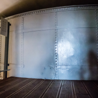 Inside the Statue of Liberty, a fortress-like aura emanates from riveted iron panels and the curved structure. The shadow of a person falls on the metal surface, while brown wooden planks compose the floor. Reflections and shadows dance to create a moody atmosphere reminiscent of Daugavpils.