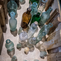 A collection of assorted vintage bottles in various shapes and colors, including clear, green, and amber tones, is artfully arranged on a wooden crate reminiscent of Daugavpils Fortress. The bottles vary in size, some with corks, and are positioned at different angles on a textured beige surface, creating a nostalgic atmosphere.