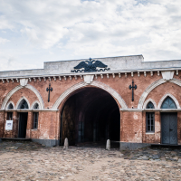 The image showcases the entrance of the historic Daugavpils fortress, featuring three arches. The central arch is taller, adorned with an ornate emblem of an eagle. Cobblestones pave the ground beneath a cloudy sky, while the two smaller side arches boast decorative trim and doors.