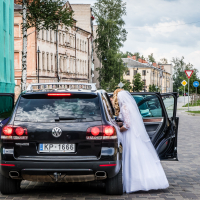 A bride in a white wedding dress leans into the backseat of a black Volkswagen SUV with a "KP-1666" license plate. The car, parked on a cobblestone street, is near the historic Daugavpils fortress, surrounded by old buildings and trees on a cloudy day with patches of blue sky.