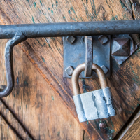 A large, weathered metal lock secures an old wooden door with visible grain and diagonal patterns, reminiscent of a fortress. The door features a sturdy metal latch and hinges with prominent rivets. The tarnished lock highlights its age against the rustic, textured surface of the Daugavpils-crafted portal.