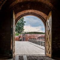 View through a brick archway with wooden doors at the historic Daugavpils Fortress in Latvia, leading to an outdoor scene with a wooden bridge. The sky is cloudy, and there are green grass and trees in the background. A person is visible in the distance to the left, near the bridge.