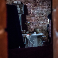 A dimly lit room in Daugavpils fortress features a table draped with a lace cloth, holding a brass candelabra and a few items. The brick walls are adorned with vintage black-and-white photographs. Dark clothing hangs on the left, partially obscuring the view. The ambiance is nostalgic and mysterious.