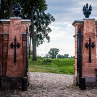 A historic site greets visitors with a brick entryway of tall red walls, adorned by black ornamental gates and large decorative hinges. The cobblestone path guides you to grassy areas with trees. With a cloudy sky above revealing patches of blue, the scene evokes the timeless charm of a fortress.