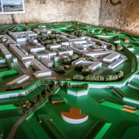 A detailed architectural model showcases a planned urban area featuring white building structures akin to a modern fortress, surrounded by a lush green landscape with small trees. Set on a table in a dimly lit room with textured walls, it evokes the charm of Latvia's historic city, Daugavpils, while photographs linger in the background.