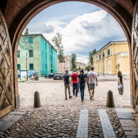 A group of five people walk toward a cobblestone street viewed through a brick archway at the Daugavpils fortress. Two large wooden doors are open, revealing colorful buildings beyond—one green and the other yellow. The sky is partly cloudy, and bollards line the pathway.