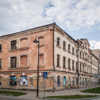 An old, dilapidated building with peeling paint and boarded windows stands at a street corner in Daugavpils. Large murals adorn its lower facade, echoing the historic charm of a fortress. A cloudy sky looms above as a cobblestone path leads past, while streetlamps and trees line the sidewalk.