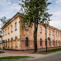 A weathered, two-story, beige building with fortress-like arched windows stands at the corner of a street in Daugavpils, surrounded by green trees. The sidewalk is lined with traffic signs, and the sky is partly cloudy. The architecture suggests historical significance.