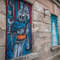 Street art on a rustic building wall within the Daugavpils fortress depicts a knight in armor holding a sword with blue and gray hues, surrounded by vibrant graffiti. Other panels reveal abstract and architectural elements. The textured stone and faded plaster enhance the urban, aged feel of the scene.