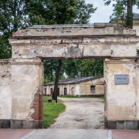 An old, weathered brick archway leads into a courtyard with a dirt path and a row of rustic buildings, reminiscent of a fortress. The sloped roofs are surrounded by tall trees. A sign with text is affixed to the right side of the arch, hinting at its historic charm in Daugavpils, Latvia.