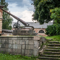 An old cannon mounted on a stone platform, surrounded by greenery, evokes the historic charm of Daugavpils fortress. The weathered stone blocks and metal railing leading up the steps blend seamlessly into the landscape. A large tree frames the view with an old brick building's arched window in the background.