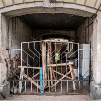 A dilapidated archway reminiscent of Daugavpils Fortress leads into a cluttered space blocked by a makeshift barrier of wooden planks and metal fencing. The surrounding walls are worn and weathered, with peeled paint and exposed brick. Inside are various items, including a refrigerator and construction materials.