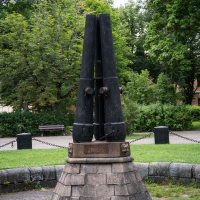 A monument with two tall, black, cylindrical structures stands on a stone base in a park reminiscent of the Daugavpils fortress. Surrounding greenery includes trees and grass, with a bench in the background. Chain barriers encircle the monument, marking its space.