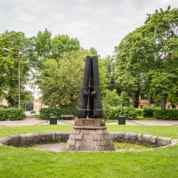 A stone pedestal with three black, vertical obelisks stands amid a circular stone enclosure, reminiscent of the historic Daugavpils fortress. Surrounded by lush green grass and tall trees, the sculpture rests under a cloudy sky with park pathways visible in the background.