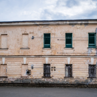 A two-story, weathered building stands within the historic Daugavpils fortress. Its faded beige paint and boarded-up windows reveal its age, with cracks and peeling paint adding character. Bars on some ground-floor windows expose patches of brick, set against a backdrop of greenery under a cloudy sky.