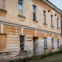 The old, two-story building with a faded orange facade and boarded-up windows stands like a forgotten sentinel near the Daugavpils Fortress. Its lower level is bricked up with overgrown grass, while the intact upper windows catch the muted light of an overcast sky, whispering stories to curious tourists.