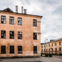 An old, weathered building with faded orange walls and multiple windows stands on a corner, reminiscent of the historical decay found in the fortresses of Daugavpils. A streetlight and paved walkway are in the foreground, while another rundown building is visible under a cloudy sky.