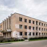 An old, worn three-story building reminiscent of a forgotten fortress stands empty in Daugavpils. Peeling paint and damaged brickwork with plants growing from cracks add to its eerie charm. Under a cloudy sky, it looms over the quiet street below. A partially visible warning sign hints at past significance.