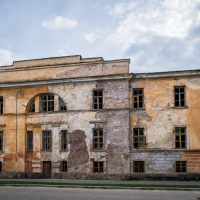 An old, abandoned building with faded yellow and orange plaster stands amid the historical charm of Daugavpils. Peeling paint, broken windows, and exposed brickwork show signs of decay against a cloudy sky. A narrow street runs along its front, reminiscent of the nearby fortress's enduring spirit.