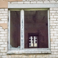 A cracked glass window on a weathered brick wall, reminiscent of a fortress in Daugavpils, Latvia. Both windows have peeling paint, creating a sense of decay. The interior is dark and empty, adding to the abandoned atmosphere. The unevenly colored bricks enhance the aged look.