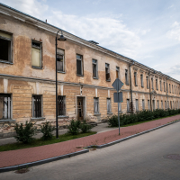 An old, weathered two-story building with peeling paint and several windows, some missing glass or blinds, stretches along an empty street near Daugavpils Fortress. The sidewalk is lined with small shrubs, and the sky is overcast with patches of blue. Two street signs are visible in the foreground.