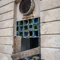 A damaged window with broken glass blocks sits on the worn brick wall of Daugavpils fortress, topped by a large circular vent. A wooden plank obstructs part of the opening, where some glass blocks are missing, revealing an interior space beyond. Peeling paint adds to the fortress's weathered charm.
