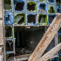 A dilapidated wall with missing and broken glass blocks reveals a dark, empty interior reminiscent of the storied Daugavpils Fortress. The glass blocks, in varying shades of blue, green, and clear, are surrounded by chipped concrete. A large wooden plank diagonally covers part of the opening, enhancing the scene's abandoned feel.