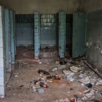A deteriorated public bathroom at the Daugavpils fortress features broken stalls and missing doors. The floor is littered with debris, including shattered tiles and discarded bottles. Cracked blue tiles line the walls, while faucets are missing from the sinks, leaving exposed pipes in this neglected atmosphere.