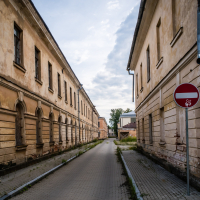 Narrow cobblestone street within Daugavpils Fortress, flanked by two historic, weathered buildings with arched windows and faded plaster. A "no entry" sign on the right and a triangular traffic sign on the left. The overcast sky casts a moody atmosphere over this urban scene.