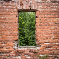 A rectangular window opening in an old, weathered red brick wall of the Daugavpils Fortress reveals dense green foliage outside. The uneven and slightly damaged bricks add a rustic charm. Wild plants grow at the base, contrasting with the vibrant greenery visible through the window.