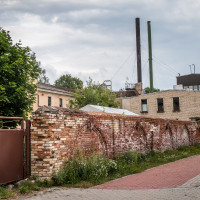 An industrial area with a brick wall and overgrown plants evokes the historic charm of Daugavpils Fortress. Behind the wall, two tall chimneys rise above several beige buildings. A closed metal gate stands on the left, while a pedestrian pathway curves alongside. The sky is cloudy.