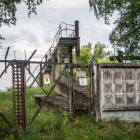 A rusted metal fence with warning signs stands in front of a concrete platform with a metal staircase leading up, hinting at the historic allure of Daugavpils Fortress. The area, enclosed by barbed wire, reveals trees and grass under a cloudy sky—a unique tourism spot steeped in mystery and history.