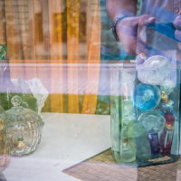 A glass jar filled with colorful bottles and ornaments sits on a window ledge alongside a lidded glass container. The window reflects an arm taking a photo, capturing the essence of Daugavpils Fortress outside, where a blurred street and building are visible. A woven mat partially covers the ledge.