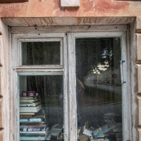 A wooden-framed window with peeling paint partially open reveals stacks of books on the sill inside. Reflections of trees and a fence are visible in the glass. The building's weathered facade, reminiscent of Daugavpils fortress, boasts textured, faded orange tones and stone detailing above the window.