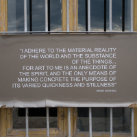 A weathered banner with a faded quote by Mark Rothko, whose roots trace back to Daugavpils, Latvia, hangs on an old, cracked wall within the fortress. The yellow-framed windows frame the message: "Art to me is an anecdote of the spirit," capturing Rothko's essence in this historic setting.
