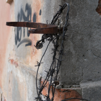 Close-up of a weathered wall at Daugavpils Fortress reveals exposed bricks and a rusty metal bracket. Coiled barbed wire loops around it, while faded spray paint graffiti hints at the fortress's age and decay seen in its concrete structure.
