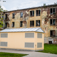 A small, modern beige building with a metal roof stands in front of the historic Daugavpils fortress, with its large, dilapidated brick walls featuring peeling paint and broken windows. The grassy area is complemented by a paved path, and leafy branches frame the scene from the upper left corner.